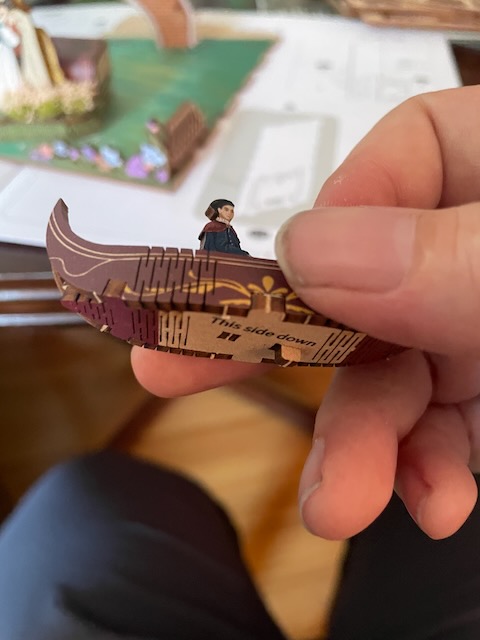 a hand holds a small 3d printed balsawood boat painted brown with gold accents. a small painted figure is sat inside. the boat is tipped away from the camera, so the text on the bottom reading 'this side down' can be seen.